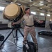 A military service member in desert camouflage uniform stands beside a large tripod-mounted radar or directed-energy equipment inside a warehouse or hangar facility. The individual appears to be gesturing or explaining something, possibly conducting a demonstration or briefing. The setting includes military equipment cases and cable reels on a concrete floor.