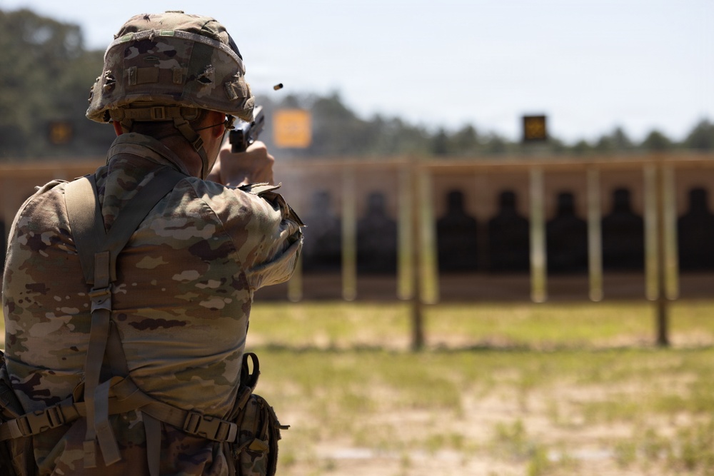 A soldier in full camouflage uniform and combat helmet is firing a pistol at a military shooting range. A spent brass casing is visibly ejecting from the firearm near the top of the frame. Multiple silhouette targets are visible downrange on a structured target berm.