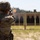 2026 US ARMY BEST SQUAD COMPETITION: Pistol Match A soldier in full camouflage uniform and combat helmet is firing a pistol at a military shooting range. A spent brass casing is visibly ejecting from the firearm near the top of the frame. Multiple silhouette targets are visible downrange on a structured target berm.
