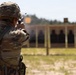 A soldier in full camouflage uniform and combat helmet is firing a pistol at a military shooting range. A spent brass casing is visibly ejecting from the firearm near the top of the frame. Multiple silhouette targets are visible downrange on a structured target berm.