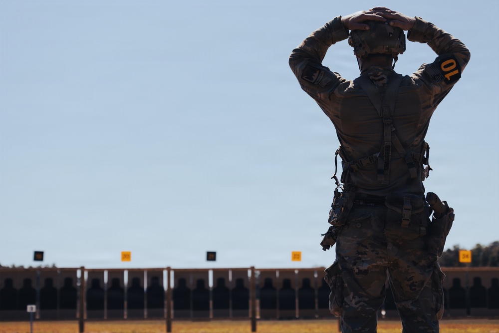 A soldier in full tactical gear stands with hands placed on the back of their head, facing a military shooting range with multiple target silhouettes. The individual is wearing multicam camouflage, a tactical vest, and carries equipment on a duty belt. The scene appears to be a live-fire range exercise or competition, indicated by the numbered lane markers visible above the target line.