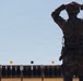 A soldier in full tactical gear stands with hands placed on the back of their head, facing a military shooting range with multiple target silhouettes. The individual is wearing multicam camouflage, a tactical vest, and carries equipment on a duty belt. The scene appears to be a live-fire range exercise or competition, indicated by the numbered lane markers visible above the target line.