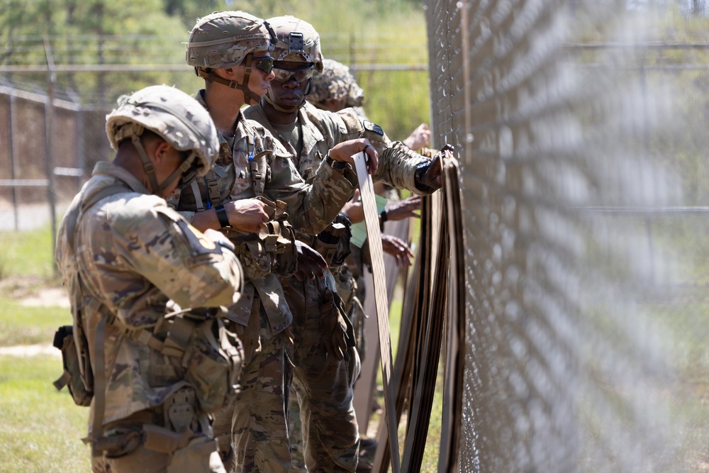 Several soldiers in military camouflage uniforms and helmets are working together near a chain-link fence, appearing to handle straps or equipment. The setting appears to be a military training facility or base, with trees and additional fencing visible in the background. The soldiers are engaged in a coordinated task involving what appears to be securing or threading material through or along the fence.