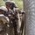 2026 US ARMY BEST SQUAD COMPETITION: Pistol Match Several soldiers in military camouflage uniforms and helmets are working together near a chain-link fence, appearing to handle straps or equipment. The setting appears to be a military training facility or base, with trees and additional fencing visible in the background. The soldiers are engaged in a coordinated task involving what appears to be securing or threading material through or along the fence.