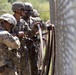 Several soldiers in military camouflage uniforms and helmets are working together near a chain-link fence, appearing to handle straps or equipment. The setting appears to be a military training facility or base, with trees and additional fencing visible in the background. The soldiers are engaged in a coordinated task involving what appears to be securing or threading material through or along the fence.
