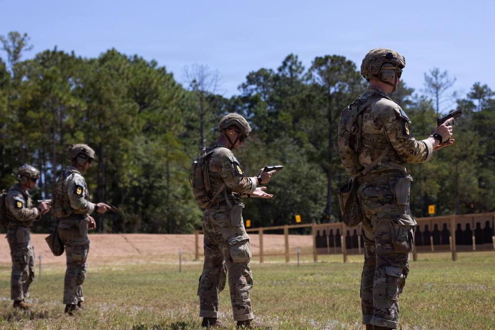 Three military personnel in full combat gear are standing at an outdoor shooting range, each handling pistols in what appears to be a training or qualification exercise. The soldiers are wearing MultiCam camouflage uniforms, tactical helmets, and body armor, and are positioned in a line facing downrange toward silhouette targets. The setting is a well-maintained open-air range surrounded by pine trees under clear skies.
