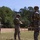 2026 US ARMY BEST SQUAD COMPETITION: Pistol Match Three military personnel in full combat gear are standing at an outdoor shooting range, each handling pistols in what appears to be a training or qualification exercise. The soldiers are wearing MultiCam camouflage uniforms, tactical helmets, and body armor, and are positioned in a line facing downrange toward silhouette targets. The setting is a well-maintained open-air range surrounded by pine trees under clear skies.