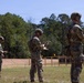 Three military personnel in full combat gear are standing at an outdoor shooting range, each handling pistols in what appears to be a training or qualification exercise. The soldiers are wearing MultiCam camouflage uniforms, tactical helmets, and body armor, and are positioned in a line facing downrange toward silhouette targets. The setting is a well-maintained open-air range surrounded by pine trees under clear skies.