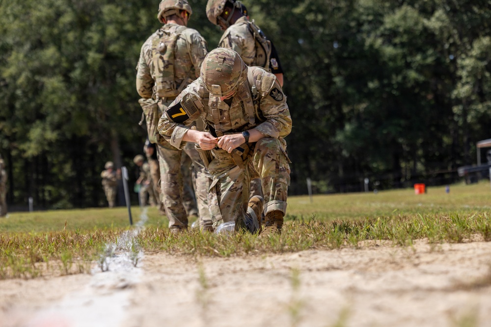 A soldier in MultiCam/OCP camouflage uniform is kneeling on a grassy field, appearing to handle a small object near the ground with what looks like a lit fuse or smoke trail visible. The soldier wears a helmet, tactical vest, and a black armband with the number '7', suggesting a competition or training event. Additional soldiers in similar uniforms are visible in the background observing the activity.