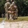 2026 US ARMY BEST SQUAD COMPETITION: Pistol Match A soldier in MultiCam/OCP camouflage uniform is kneeling on a grassy field, appearing to handle a small object near the ground with what looks like a lit fuse or smoke trail visible. The soldier wears a helmet, tactical vest, and a black armband with the number '7', suggesting a competition or training event. Additional soldiers in similar uniforms are visible in the background observing the activity.