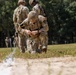 A soldier in MultiCam/OCP camouflage uniform is kneeling on a grassy field, appearing to handle a small object near the ground with what looks like a lit fuse or smoke trail visible. The soldier wears a helmet, tactical vest, and a black armband with the number '7', suggesting a competition or training event. Additional soldiers in similar uniforms are visible in the background observing the activity.