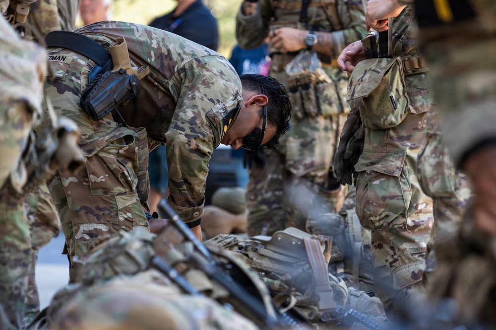 A group of military personnel in MultiCam/OCP camouflage uniforms are gathered around equipment laid out on the ground. One soldier is bent over inspecting or organizing gear, including what appears to be a rifle and tactical equipment. The scene appears to be a pre-mission equipment check or gear inspection.