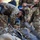 2026 US ARMY BEST SQUAD COMPETITION: Pistol Match A group of military personnel in MultiCam/OCP camouflage uniforms are gathered around equipment laid out on the ground. One soldier is bent over inspecting or organizing gear, including what appears to be a rifle and tactical equipment. The scene appears to be a pre-mission equipment check or gear inspection.