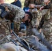 A group of military personnel in MultiCam/OCP camouflage uniforms are gathered around equipment laid out on the ground. One soldier is bent over inspecting or organizing gear, including what appears to be a rifle and tactical equipment. The scene appears to be a pre-mission equipment check or gear inspection.