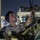 A U.S. Marine in desert MARPAT camouflage uniform is working on military vehicle equipment at night, appearing to adjust or install an antenna or similar component. The scene takes place in what appears to be a port or staging area, with industrial structures and lights visible in the background. At least one other service member is visible in the background.