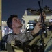 A U.S. Marine in desert MARPAT camouflage uniform is working on military vehicle equipment at night, appearing to adjust or install an antenna or similar component. The scene takes place in what appears to be a port or staging area, with industrial structures and lights visible in the background. At least one other service member is visible in the background.