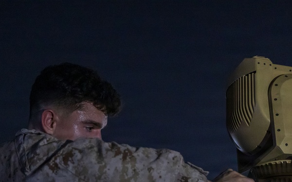 A military service member in desert camouflage uniform is working on what appears to be a weapons or sensor system at night. The individual is closely inspecting or adjusting a turret-mounted optical/sensor unit, likely a targeting or surveillance system. The setting appears to be an outdoor military installation or airfield based on background lighting.