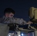 A military service member in desert camouflage uniform is working on what appears to be a weapons or sensor system at night. The individual is closely inspecting or adjusting a turret-mounted optical/sensor unit, likely a targeting or surveillance system. The setting appears to be an outdoor military installation or airfield based on background lighting.