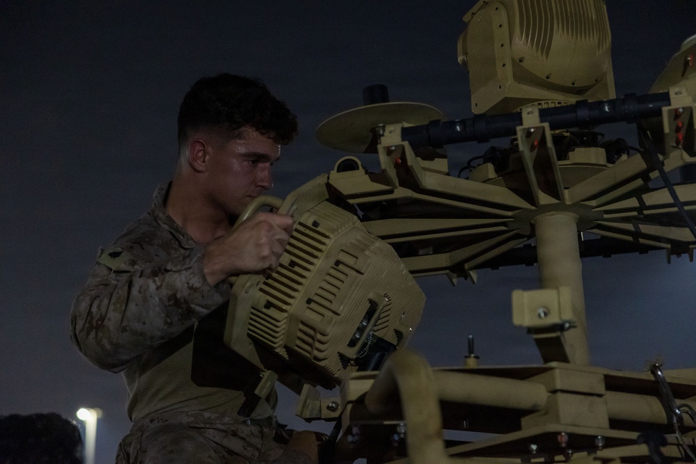 A military service member in camouflage uniform is working on a piece of tan/desert-colored military equipment at night or dusk. The individual appears to be performing maintenance or assembly on what looks like a radar or sensor system with multiple components. The scene is illuminated by ambient artificial lighting against a dark sky.