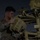 A military service member in camouflage uniform is working on a piece of tan/desert-colored military equipment at night or dusk. The individual appears to be performing maintenance or assembly on what looks like a radar or sensor system with multiple components. The scene is illuminated by ambient artificial lighting against a dark sky.