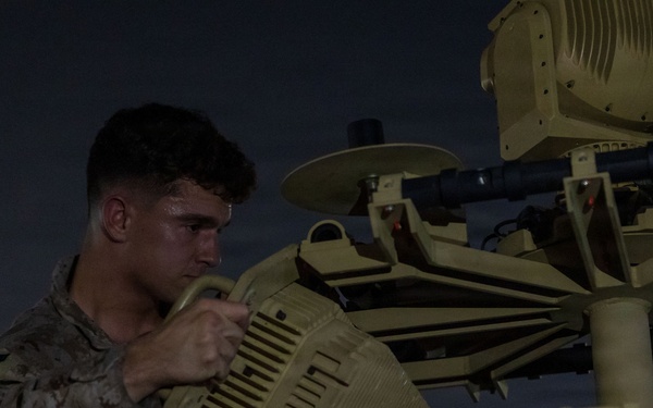 A military service member in camouflage uniform is working on a piece of tan/desert-colored military equipment at night or dusk. The individual appears to be performing maintenance or assembly on what looks like a radar or sensor system with multiple components. The scene is illuminated by ambient artificial lighting against a dark sky.