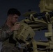 A military service member in camouflage uniform is working on a piece of tan/desert-colored military equipment at night or dusk. The individual appears to be performing maintenance or assembly on what looks like a radar or sensor system with multiple components. The scene is illuminated by ambient artificial lighting against a dark sky.