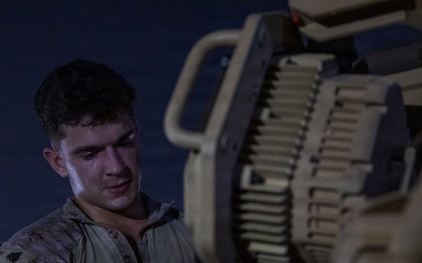 A young male service member in U.S. Marine Corps MARPAT desert camouflage uniform is working on military equipment at night. He appears to be performing maintenance or loading operations on what appears to be a crew-served weapon system or vehicle-mounted equipment. The scene is illuminated by artificial light against a dark nighttime background.