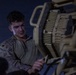 A young male service member in U.S. Marine Corps MARPAT desert camouflage uniform is working on military equipment at night. He appears to be performing maintenance or loading operations on what appears to be a crew-served weapon system or vehicle-mounted equipment. The scene is illuminated by artificial light against a dark nighttime background.
