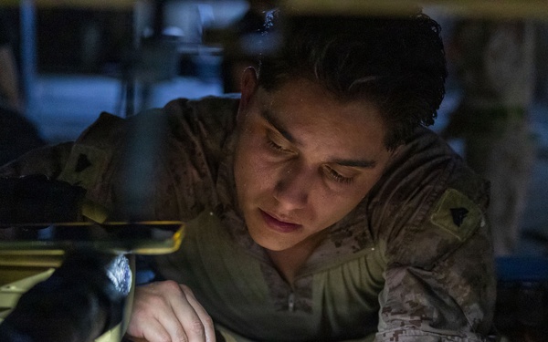 A military service member in desert MARPAT camouflage uniform is closely examining or working on a piece of equipment, likely a helmet or tactical gear with cables and attachments. The individual is leaning over a work surface in what appears to be an indoor, dimly lit workspace. The photo is taken at close range with dramatic lighting highlighting the subject's focused expression.