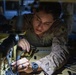 A military service member in desert MARPAT camouflage uniform is closely examining or working on a piece of equipment, likely a helmet or tactical gear with cables and attachments. The individual is leaning over a work surface in what appears to be an indoor, dimly lit workspace. The photo is taken at close range with dramatic lighting highlighting the subject's focused expression.