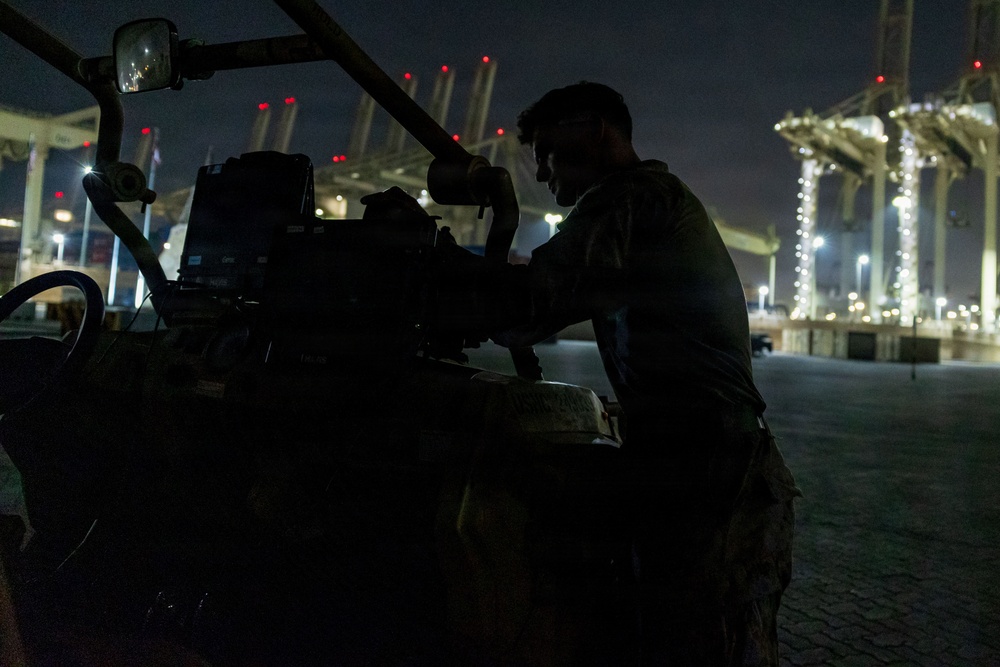 A person in military-style clothing works on heavy equipment or machinery at night in what appears to be a port or industrial facility. The scene is predominantly dark with the subject silhouetted against the illuminated background. Large port cranes or gantry structures are visible in the background, lit with red warning lights and white work lights.