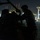 A person in military-style clothing works on heavy equipment or machinery at night in what appears to be a port or industrial facility. The scene is predominantly dark with the subject silhouetted against the illuminated background. Large port cranes or gantry structures are visible in the background, lit with red warning lights and white work lights.