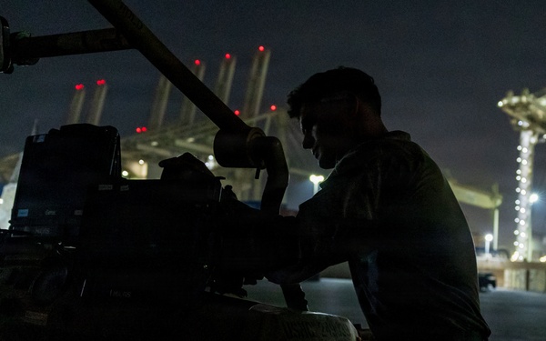 A person in military-style clothing works on heavy equipment or machinery at night in what appears to be a port or industrial facility. The scene is predominantly dark with the subject silhouetted against the illuminated background. Large port cranes or gantry structures are visible in the background, lit with red warning lights and white work lights.
