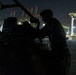 A person in military-style clothing works on heavy equipment or machinery at night in what appears to be a port or industrial facility. The scene is predominantly dark with the subject silhouetted against the illuminated background. Large port cranes or gantry structures are visible in the background, lit with red warning lights and white work lights.