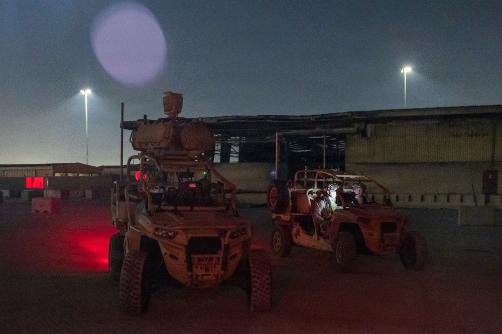 A nighttime scene at what appears to be a military installation or forward operating base, showing two desert-tan tactical military vehicles parked on a dirt surface. The left vehicle is heavily equipped with what appears to be a sensor or weapons system mounted on top. The scene is illuminated by overhead floodlights and a red light emanating from the left vehicle.