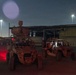 A nighttime scene at what appears to be a military installation or forward operating base, showing two desert-tan tactical military vehicles parked on a dirt surface. The left vehicle is heavily equipped with what appears to be a sensor or weapons system mounted on top. The scene is illuminated by overhead floodlights and a red light emanating from the left vehicle.