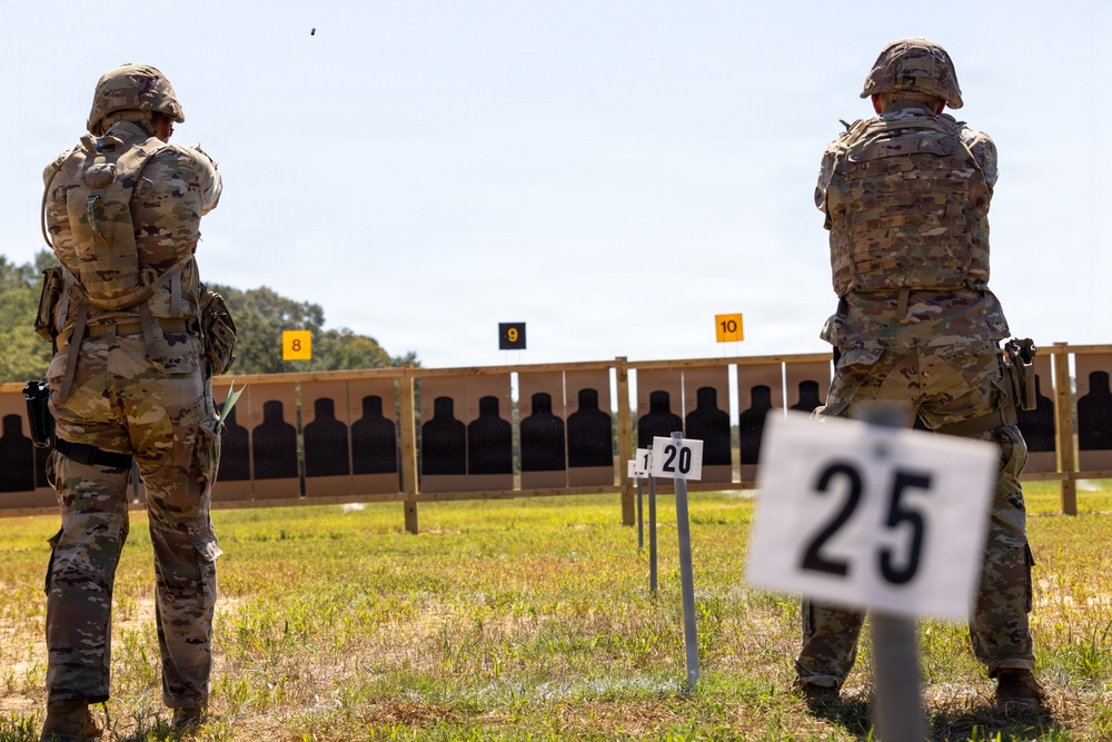 Two military personnel in full combat gear and camouflage uniforms are engaged in a live-fire shooting exercise at an outdoor firing range. The shooter on the left is actively firing a pistol, with a spent casing visibly ejecting in the air. Numbered distance markers (20 and 25 yards) and lane markers (8, 9, 10) are visible, with a row of silhouette targets downrange.