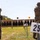 2026 US ARMY BEST SQUAD COMPETITION: Pistol Match Two military personnel in full combat gear and camouflage uniforms are engaged in a live-fire shooting exercise at an outdoor firing range. The shooter on the left is actively firing a pistol, with a spent casing visibly ejecting in the air. Numbered distance markers (20 and 25 yards) and lane markers (8, 9, 10) are visible, with a row of silhouette targets downrange.