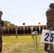 Two military personnel in full combat gear and camouflage uniforms are engaged in a live-fire shooting exercise at an outdoor firing range. The shooter on the left is actively firing a pistol, with a spent casing visibly ejecting in the air. Numbered distance markers (20 and 25 yards) and lane markers (8, 9, 10) are visible, with a row of silhouette targets downrange.