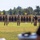 2026 US ARMY BEST SQUAD COMPETITION: Pistol Match An outdoor shooting range is depicted with multiple personnel standing downrange near numbered target bays (visible numbers 19-23). Two individuals in full tactical/military gear and one person in a dark shirt and tan pants (appearing to hold a scoring paddle) are positioned in front of silhouette paper targets mounted on wooden frames. The scene appears to be a formal marksmanship competition or qualification event.
