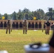 An outdoor shooting range is depicted with multiple personnel standing downrange near numbered target bays (visible numbers 19-23). Two individuals in full tactical/military gear and one person in a dark shirt and tan pants (appearing to hold a scoring paddle) are positioned in front of silhouette paper targets mounted on wooden frames. The scene appears to be a formal marksmanship competition or qualification event.