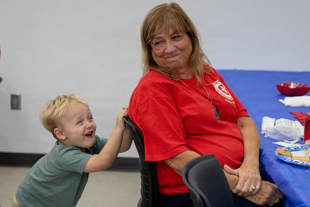 An older woman wearing a red t-shirt sits at a table with a blue tablecloth, smiling warmly at the camera. A young blonde toddler in a green t-shirt stands behind her chair, laughing and gripping the back of her chair playfully. The setting appears to be an indoor gathering or event, possibly a community or family celebration.