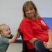 An older woman wearing a red t-shirt sits at a table with a blue tablecloth, smiling warmly at the camera. A young blonde toddler in a green t-shirt stands behind her chair, laughing and gripping the back of her chair playfully. The setting appears to be an indoor gathering or event, possibly a community or family celebration.