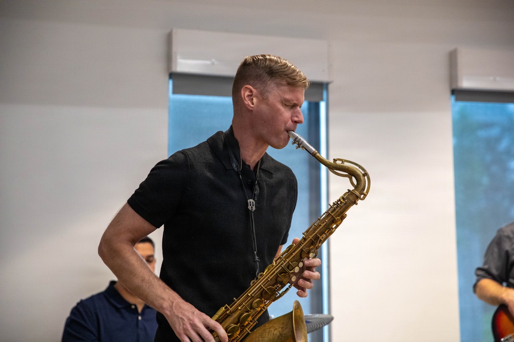 A man in a black short-sleeve polo shirt is actively playing a brass tenor saxophone in what appears to be an indoor rehearsal or performance space. He is focused on his playing with eyes directed downward toward the instrument. At least two other individuals are partially visible in the background.