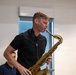 A man in a black short-sleeve polo shirt is actively playing a brass tenor saxophone in what appears to be an indoor rehearsal or performance space. He is focused on his playing with eyes directed downward toward the instrument. At least two other individuals are partially visible in the background.