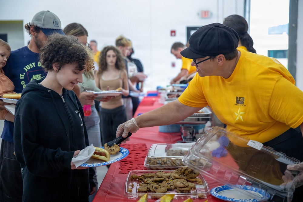A food service event where volunteers in yellow 'Gold Star Program' shirts are serving food to a line of attendees. A young person with curly hair is being served cookies and already holds a hot dog on a plate. The event appears to be a community gathering or organized meal service held in a large indoor facility.