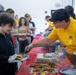A food service event where volunteers in yellow 'Gold Star Program' shirts are serving food to a line of attendees. A young person with curly hair is being served cookies and already holds a hot dog on a plate. The event appears to be a community gathering or organized meal service held in a large indoor facility.