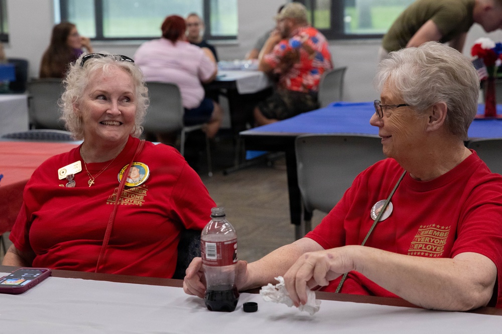 Two older women wearing matching red 'Remember Everyone Deployed' (R.E.D.) t-shirts are seated at a table, engaged in cheerful conversation. They both wear name badges and pins, suggesting participation in a veterans support or military family event held in a community hall or recreation center. The background shows additional attendees seated at tables with red and blue tablecloths.