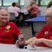 Two older women wearing matching red 'Remember Everyone Deployed' (R.E.D.) t-shirts are seated at a table, engaged in cheerful conversation. They both wear name badges and pins, suggesting participation in a veterans support or military family event held in a community hall or recreation center. The background shows additional attendees seated at tables with red and blue tablecloths.