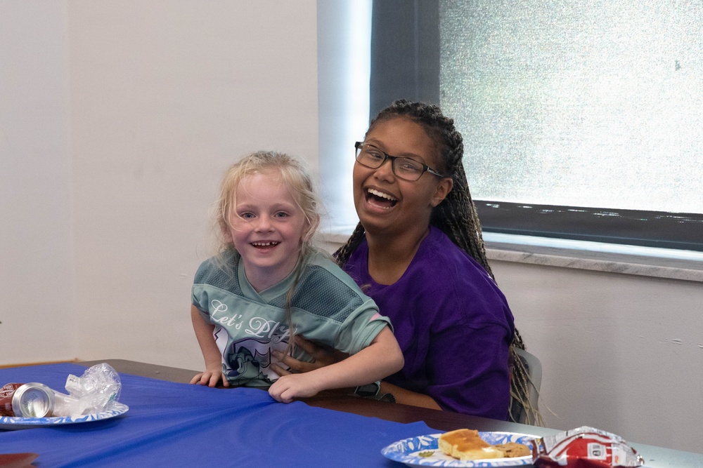 Two young girls are seated together at a table covered with a blue tablecloth, both smiling and laughing joyfully. The older girl, wearing glasses and a purple shirt, has her arm around the younger girl who is wearing a teal jersey-style shirt. The setting appears to be an indoor gathering or party with food visible on the table.